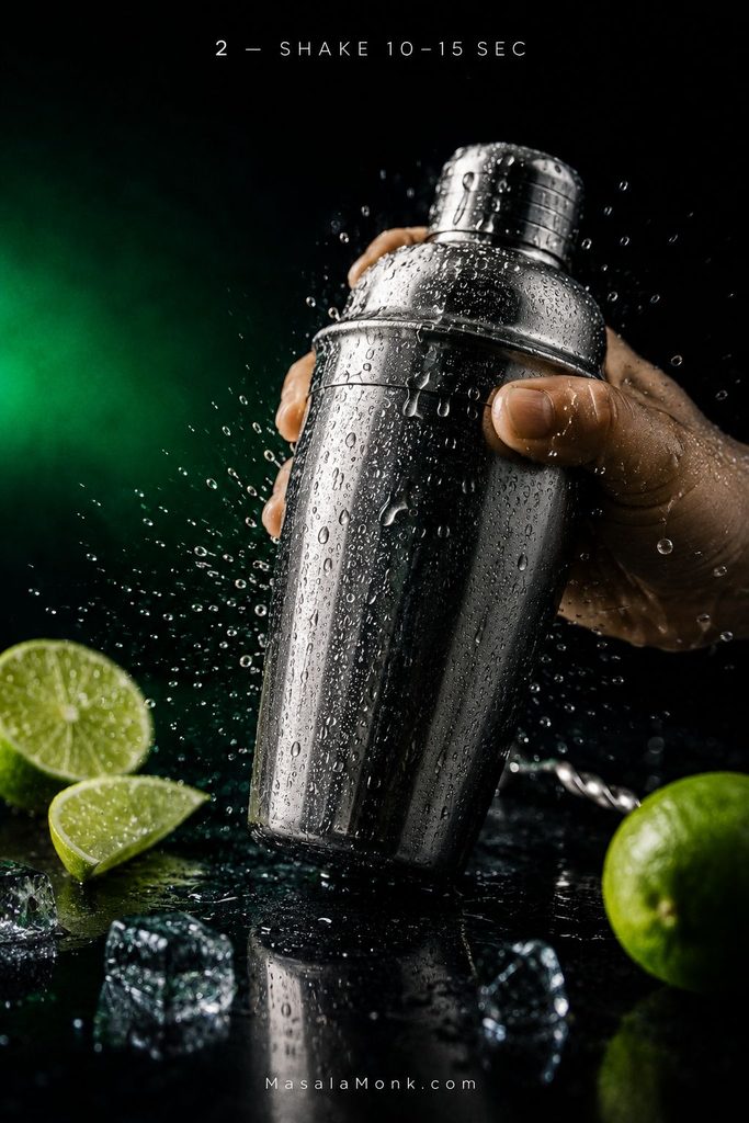 Hands shaking a condensation-covered stainless steel cocktail shaker beside fresh limes and clear ice on a dark surface.