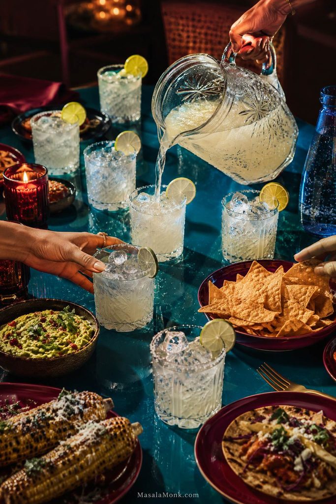 Hand pouring pale margarita mocktail base from a large crystal pitcher into glasses around a teal dinner table with chips, guacamole, grilled corn and other food.