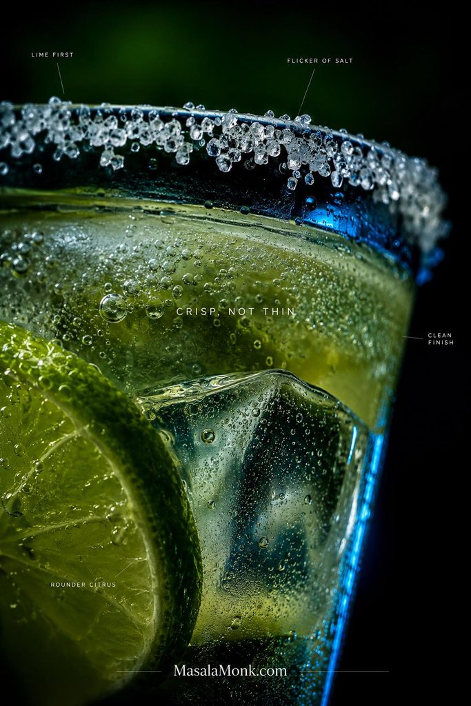 Extreme close-up of a margarita mocktail showing coarse salt crystals on the rim, lime, clear ice, condensation and tiny bubbles in pale citrus liquid.