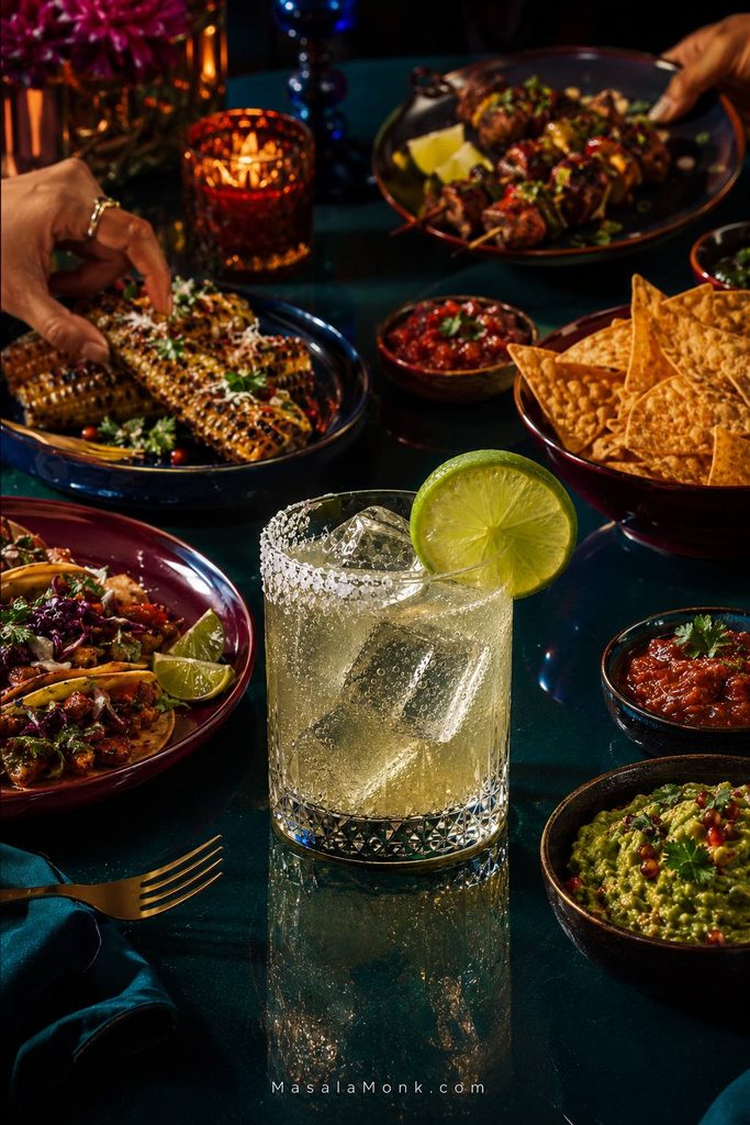Margarita mocktail on a dark teal dinner table surrounded by tacos, grilled corn, tortilla chips, salsa, guacamole and skewered grilled food, with hands reaching toward the dishes.