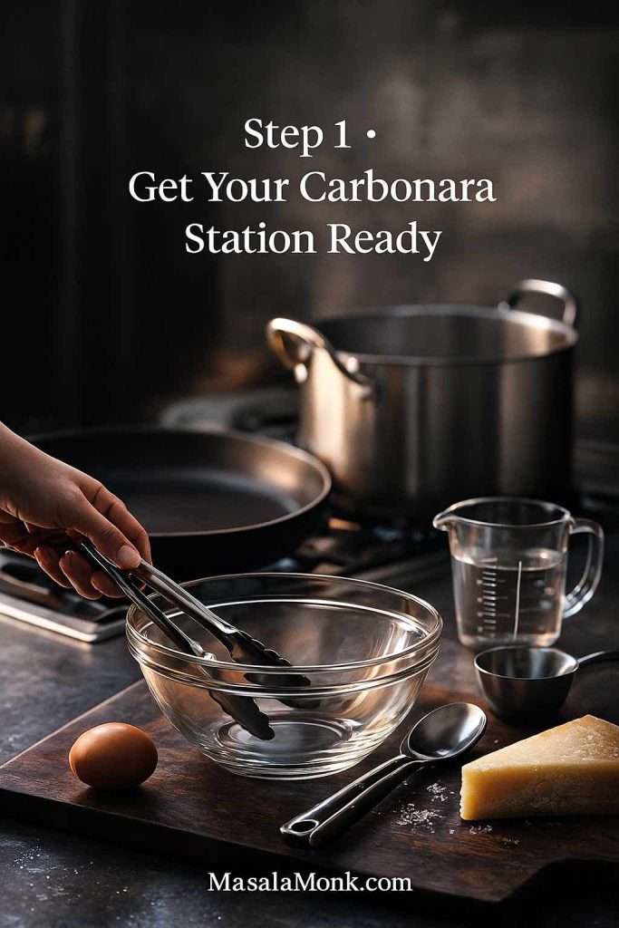 Vertical photo of a carbonara cooking station with a pot on the stove, a skillet, glass mixing bowl, tongs, measuring cup of water, an egg and a wedge of cheese on a dark counter as a hand sets the tools in place.