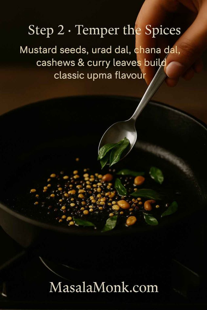Step 2 of making upma showing mustard seeds, urad dal, chana dal and cashews sizzling in oil in a black skillet while a hand adds fresh curry leaves with a spoon, illustrating the tempering stage for classic rava upma.