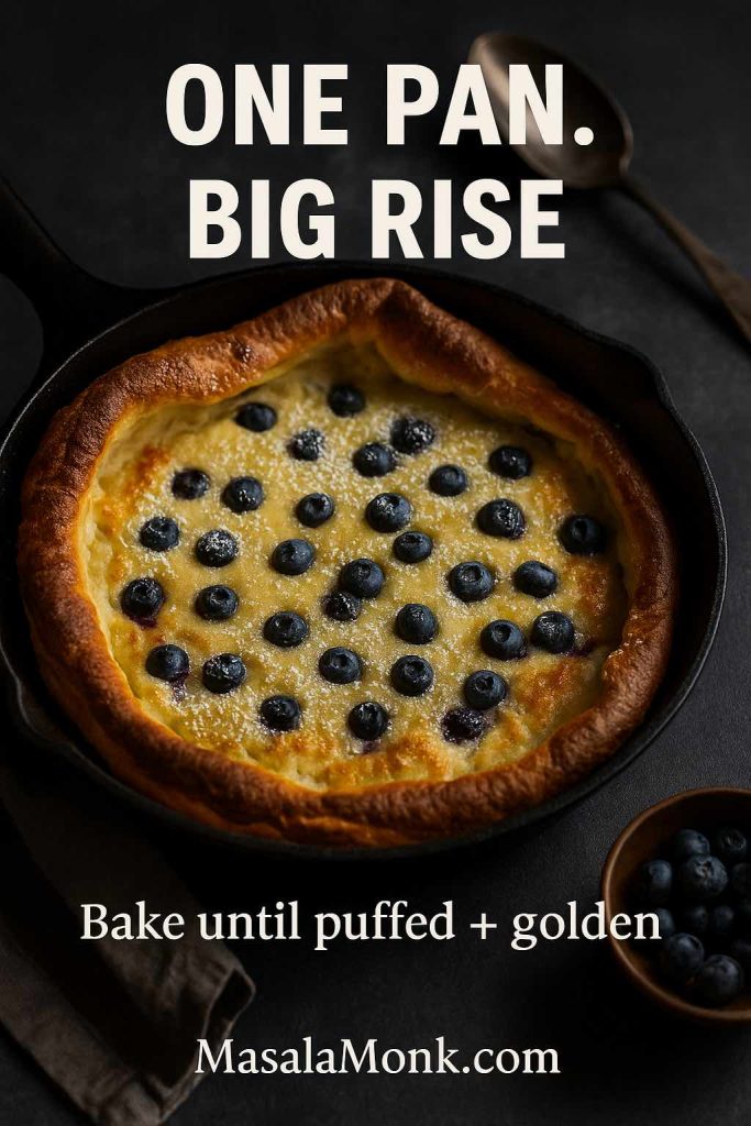 Portrait photo of a blueberry Dutch pancake baked in a cast-iron skillet with puffed golden edges and blueberries on top, featuring text overlay “One Pan. Big Rise.” and “Bake until puffed + golden,” with MasalaMonk.com in the footer.