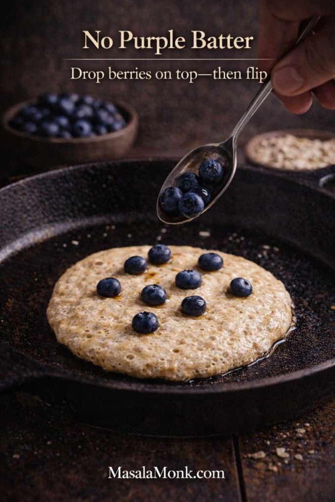 Single oat pancake cooking in a cast-iron skillet with blueberries being placed on top before flipping, with text “No Purple Batter—Drop berries on top, then flip.”