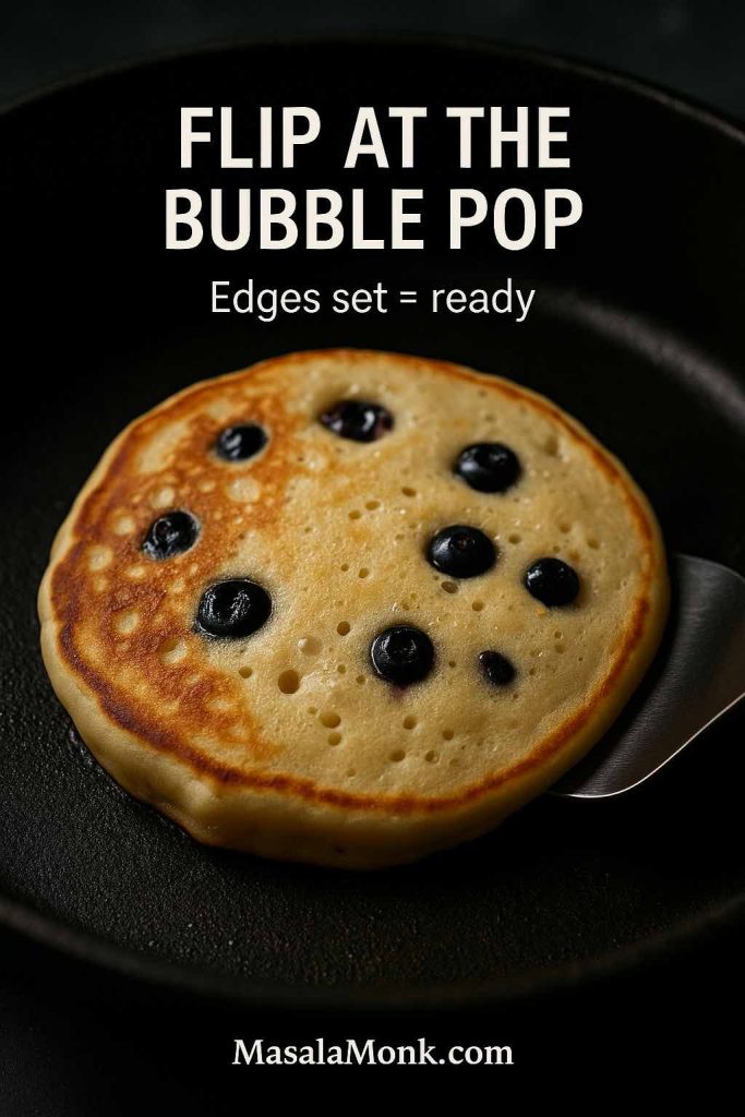 Portrait close-up of a blueberry pancake cooking in a skillet with bubbles on the surface and a spatula sliding underneath, featuring text overlay “Flip at the Bubble Pop” and “Edges set = ready,” with MasalaMonk.com in the footer.