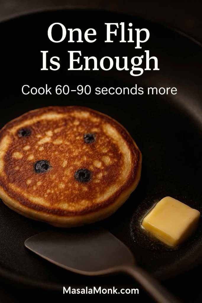 Portrait photo of a blueberry pancake in a skillet after flipping, showing an even golden-brown surface and a pat of butter melting nearby, with text overlay “One Flip Is Enough” and “Cook 60–90 seconds more,” plus MasalaMonk.com in the footer.