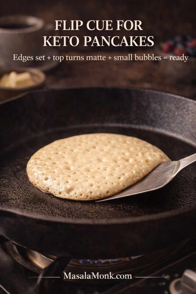 Keto pancake in a skillet with a spatula sliding underneath, showing the flip cue with set edges and small bubbles on top for fluffy low carb pancakes.