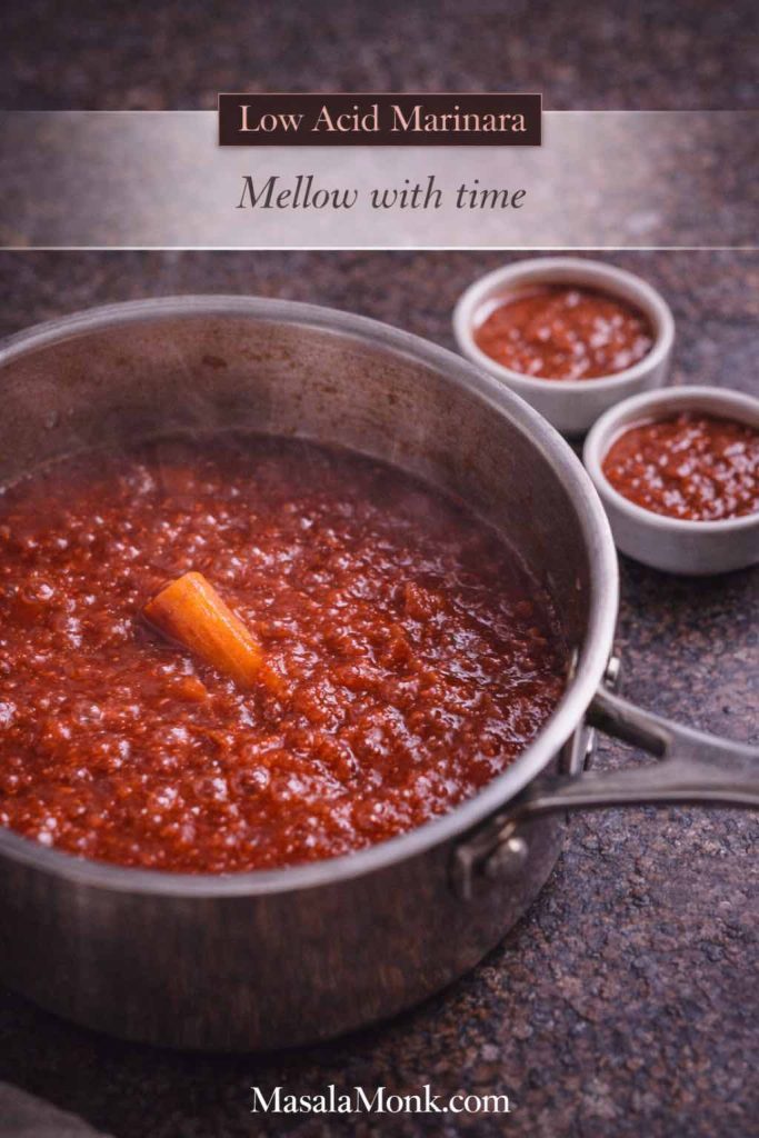 Marinara sauce gently simmering with a piece of carrot in the pot and two ramekins in the background, showing a low acid marinara method that softens sharp tomato flavor.