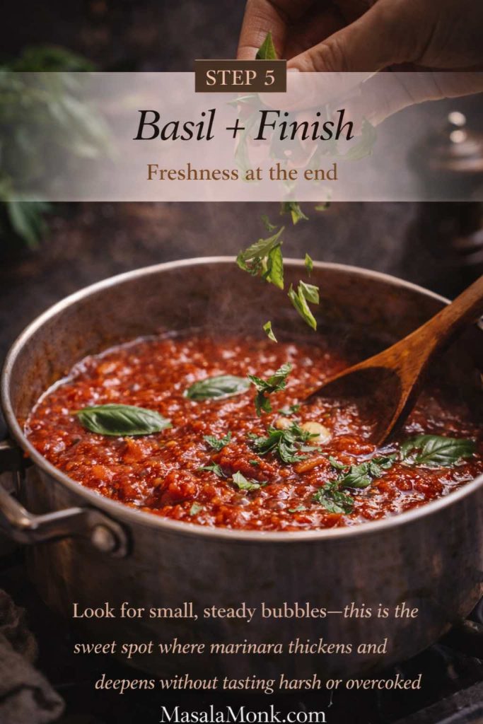 Fresh basil being torn and sprinkled into a pot of thickened marinara sauce, showing step 5 of a homemade marinara sauce recipe just before serving.