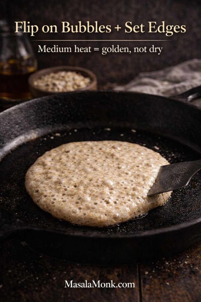 Oat pancake cooking in a cast-iron skillet with bubbles on the surface and a spatula sliding underneath, with text “Flip on Bubbles + Set Edges.”