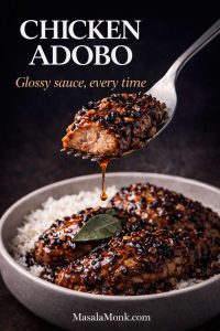A close-up fork lifts a juicy bite of chicken adobo over a bowl of rice, with a thick glossy soy-vinegar sauce dripping from the bite. The chicken is coated in dark caramelized sauce with visible garlic bits and peppercorns against a moody black background, with the text “Chicken Adobo” and “Glossy sauce, every time” plus MasalaMonk.com at the bottom.