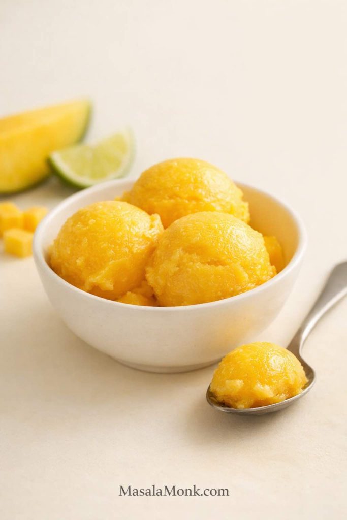 Closing hero image for a mango sorbet recipe showing three smooth scoops of bright homemade mango sorbet in a white bowl with a spoonful beside it, plus soft mango and lime accents in the background.