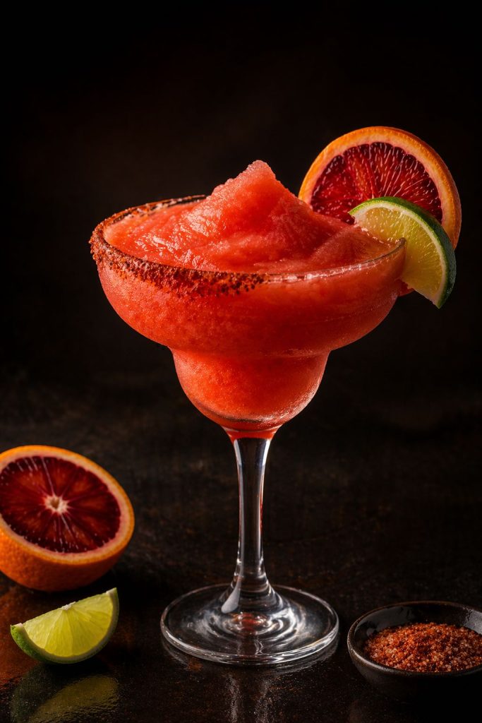 Frozen blood orange margarita in a stemmed margarita glass with a Tajín rim, blood orange slice, lime wedge, and slushy ruby-orange texture on a dark editorial background.