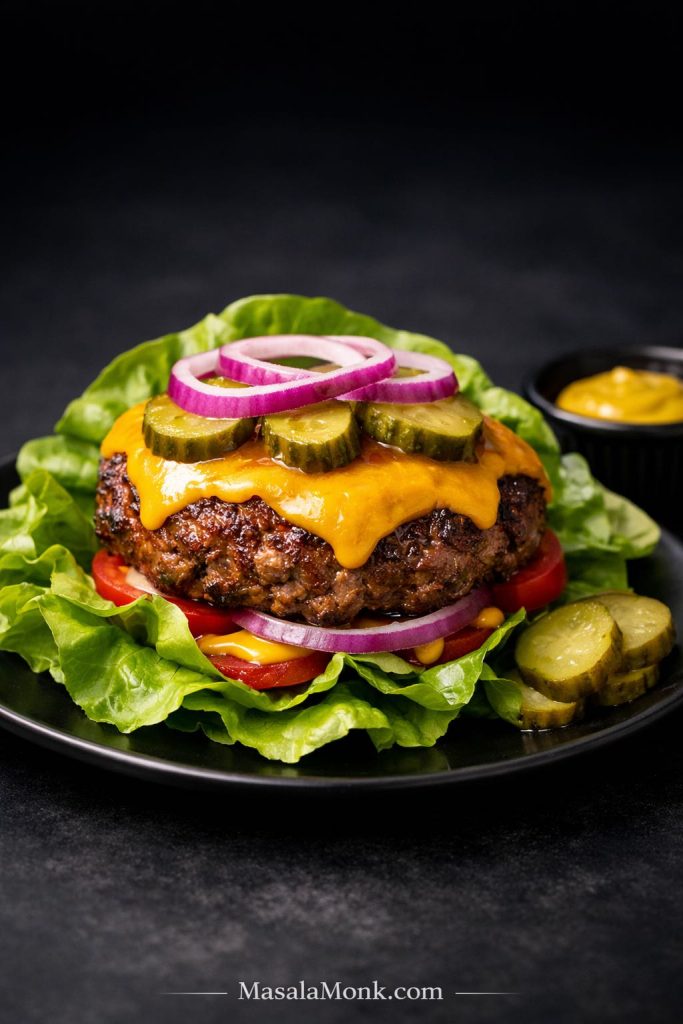 Classic bunless keto cheeseburger with melted cheddar, pickles, red onion, mustard, tomato, and lettuce leaves on a black plate.