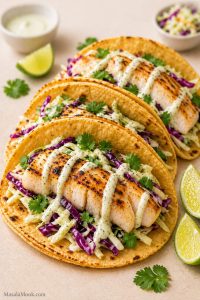 Three cod fish tacos in warm corn tortillas with cabbage slaw, lime crema, cilantro, and lime wedges.
