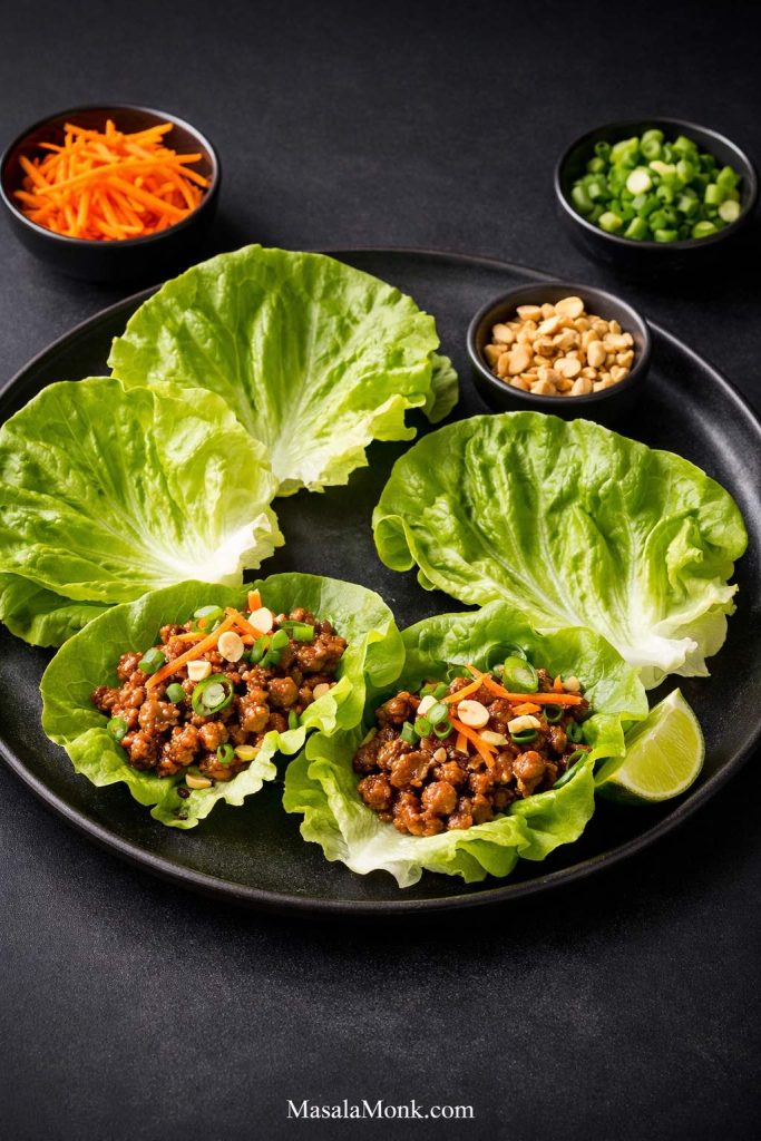 Ground pork lettuce wraps with two filled lettuce cups, extra leaves, peanuts, scallions, carrots, and lime on a black plate.