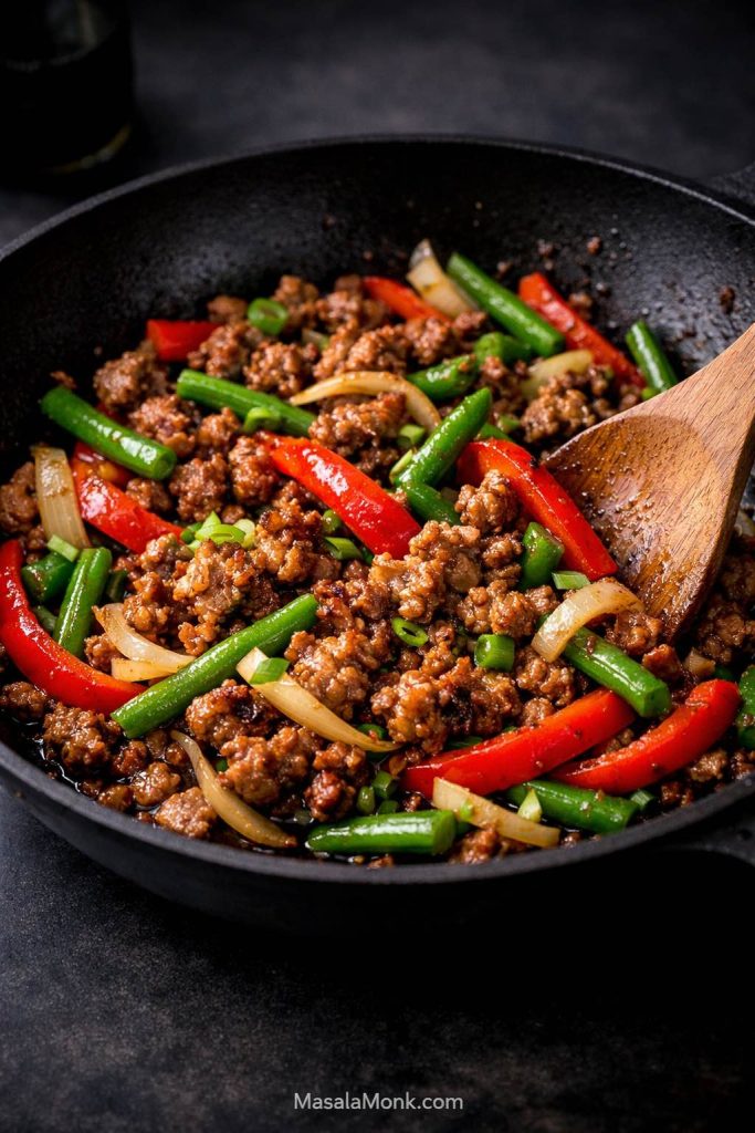 Ground pork stir fry process image showing browned pork with red peppers, green beans, onions, and a wooden spoon in a hot skillet.