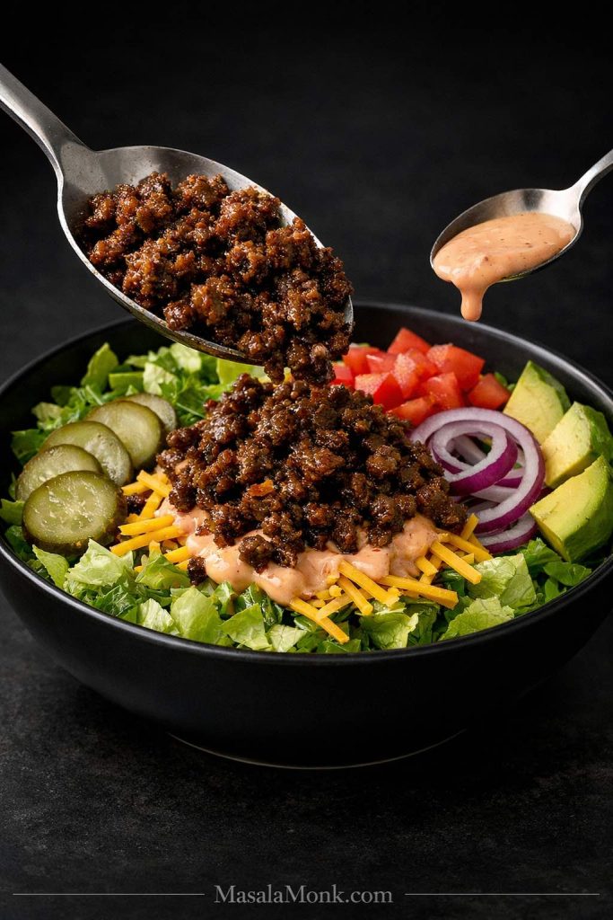 Hot browned beef and burger sauce being added to a keto burger bowl with lettuce, cheddar, pickles, red onion, tomato, and avocado.