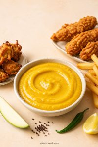 Creamy mango mustard sauce in a bowl with fries, fried chicken, pakoras, green mango, mustard seeds, chili and lemon for a quick dipping sauce.