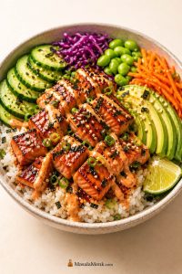 Salmon bowl recipe with pan-seared salmon bites, rice, avocado, cucumber, edamame, cabbage, carrots, sesame, scallions, lime, and creamy sauce.