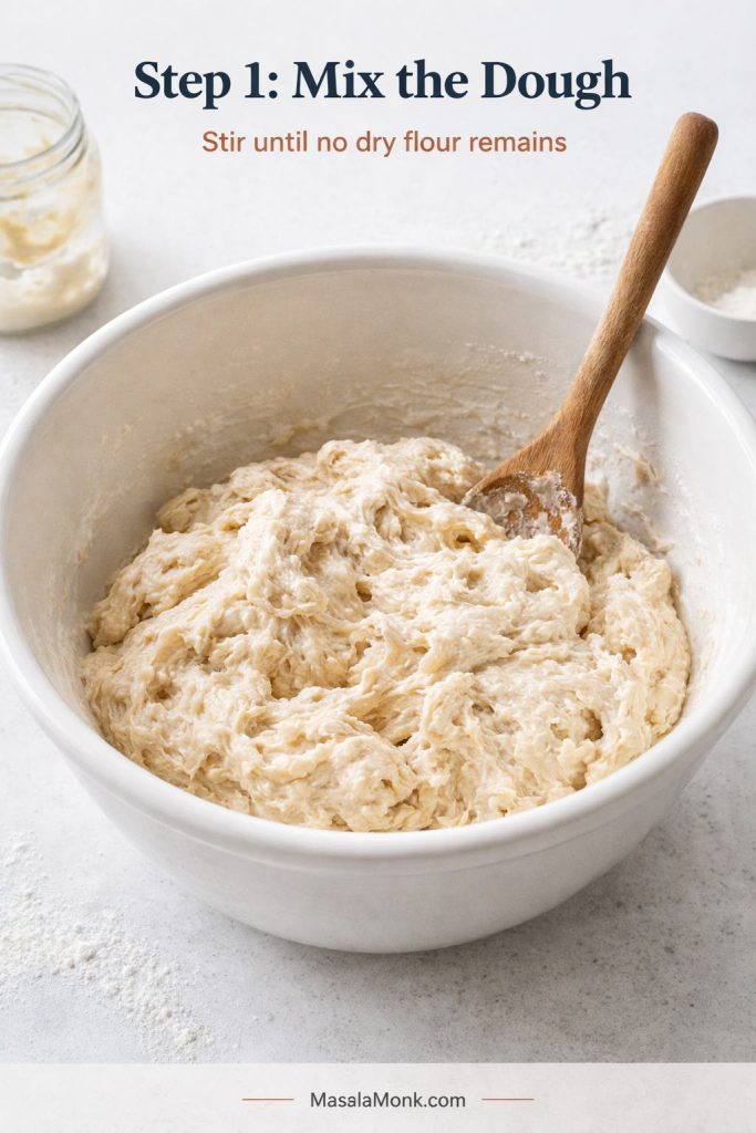 Step 1 process image for sourdough English muffins showing freshly mixed dough in a large bowl, rough and shaggy but fully combined, with no dry flour remaining and a wooden spoon resting in the dough.