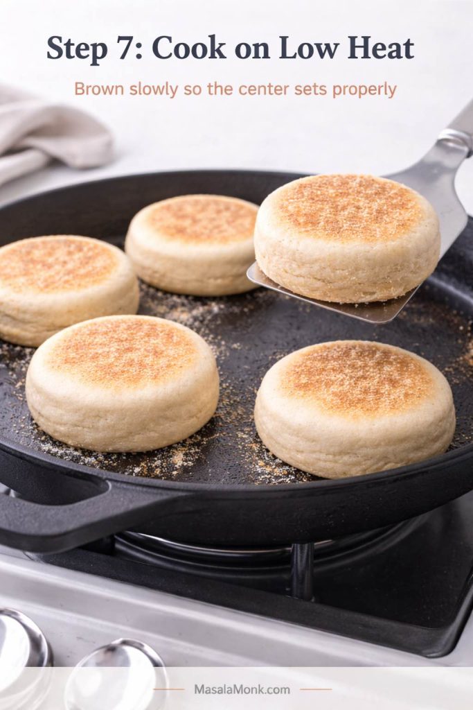 Step 7 process image for sourdough English muffins showing thick muffins cooking on a skillet over low heat, with gradual even browning and one muffin lifted on a spatula to show the underside color.