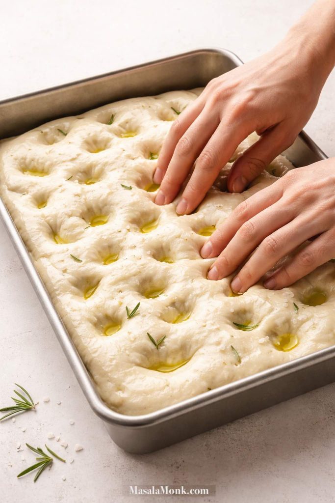 Hands dimpling proofed sourdough focaccia dough in a 9x13 metal pan with olive oil before baking.
