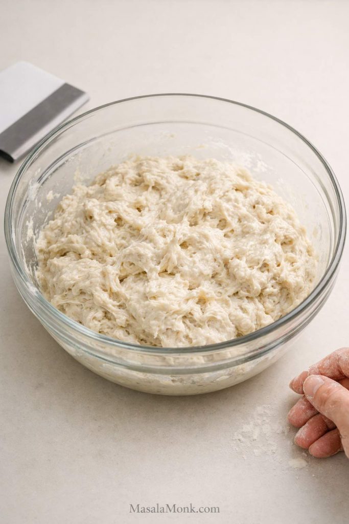 Freshly mixed sourdough focaccia dough in a glass bowl showing a rough sticky high-hydration early stage before folds.