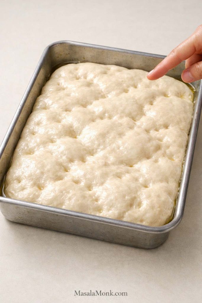 Proofed sourdough focaccia dough in a 9x13 metal pan showing a puffy airy surface ready to dimple before baking.