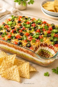 Finished 7 layer dip in a clear 9x13 glass dish with visible layers, colorful toppings, tortilla chips, and one corner lightly scooped.