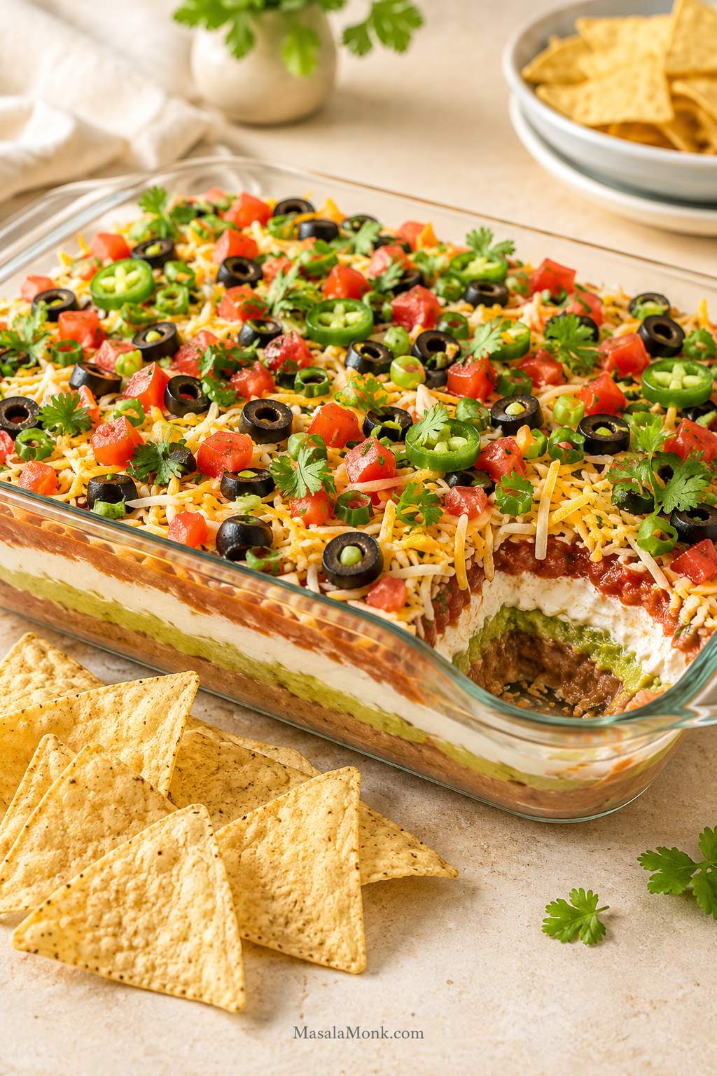 Finished 7 layer dip in a clear 9x13 glass dish with visible layers, colorful toppings, tortilla chips, and one corner lightly scooped.