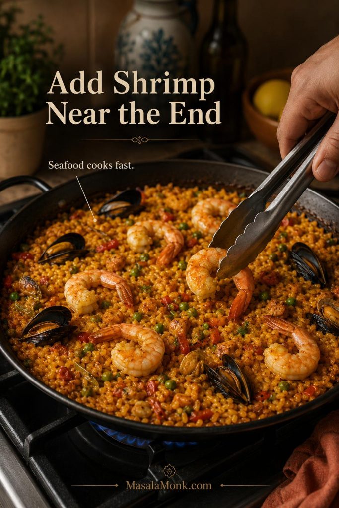 Shrimp being added near the end of cooking on top of saffron paella rice in a wide pan.