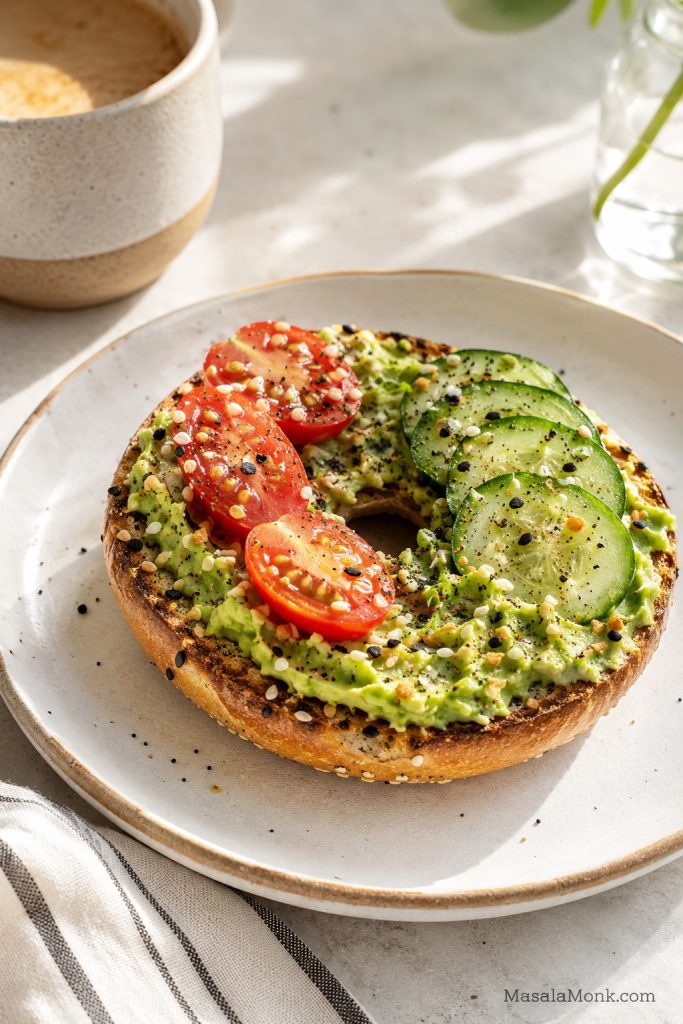 Avocado bagel toast on half a toasted bagel with seasoned avocado, cucumber, tomato, and everything seasoning.