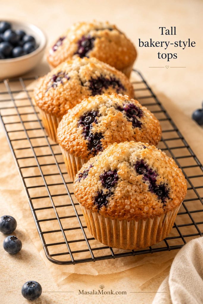Tall bakery-style blueberry muffins with golden domed tops and coarse sugar on a cooling rack.