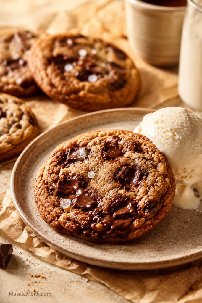 Warm brown butter chocolate chip cookie served with vanilla ice cream, melted chocolate, milk, and more cookies in the background.