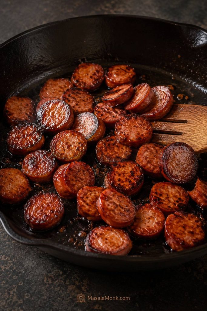 Sliced kielbasa browning in a dark skillet with seared edges, rendered fat, and a spatula.