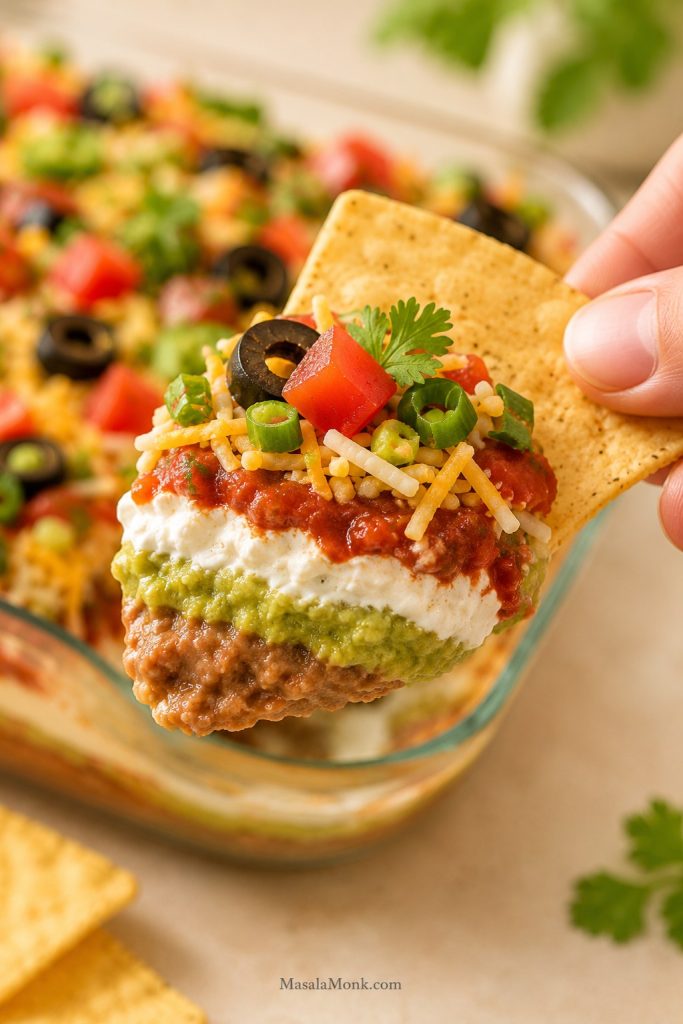 Tortilla chip lifting a clean scoop of 7 layer dip with refried beans, guacamole, sour cream, salsa, cheese, tomatoes, olives, and green onions.