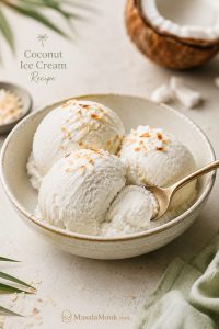 Three scoops of creamy coconut ice cream in a light ceramic bowl with toasted coconut flakes and a spoon cutting into one scoop.