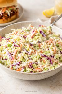 Creamy coleslaw in a bowl with shredded green cabbage, red cabbage, carrots, and a spoon lifting a serving.