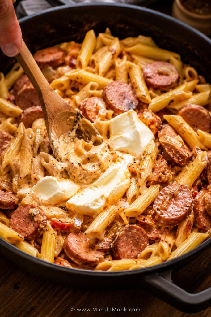 Cream cheese being stirred into kielbasa pasta in a skillet with sausage slices, pasta, and creamy sauce.