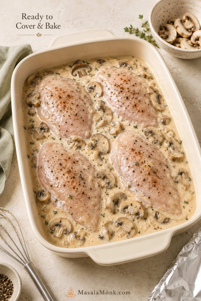 Raw chicken breasts in a baking dish with cream of mushroom sauce, sliced mushrooms, foil, and a spoon nearby.