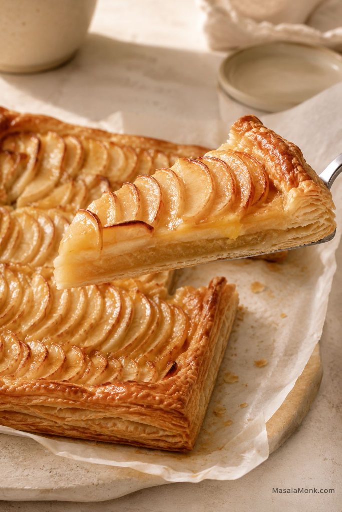 Slice of apple tart lifted from a rectangular puff pastry tart, showing glossy apples and flaky pastry layers.