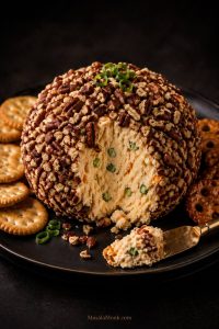 Close-up of a pecan-coated cheese ball on a dark plate with round crackers and a spreader, with a scoop taken out to show the creamy cheddar and green onion filling inside.