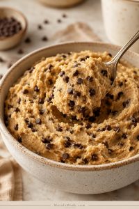 A spoon lifts soft chocolate chip edible cookie dough from a bowl, showing a creamy texture with mini chocolate chips throughout.