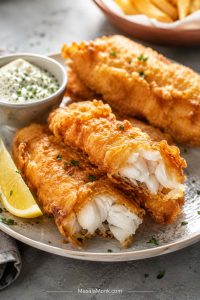 Golden battered fish pieces on a plate with one piece broken open to show flaky white fish, served with lemon and dipping sauce