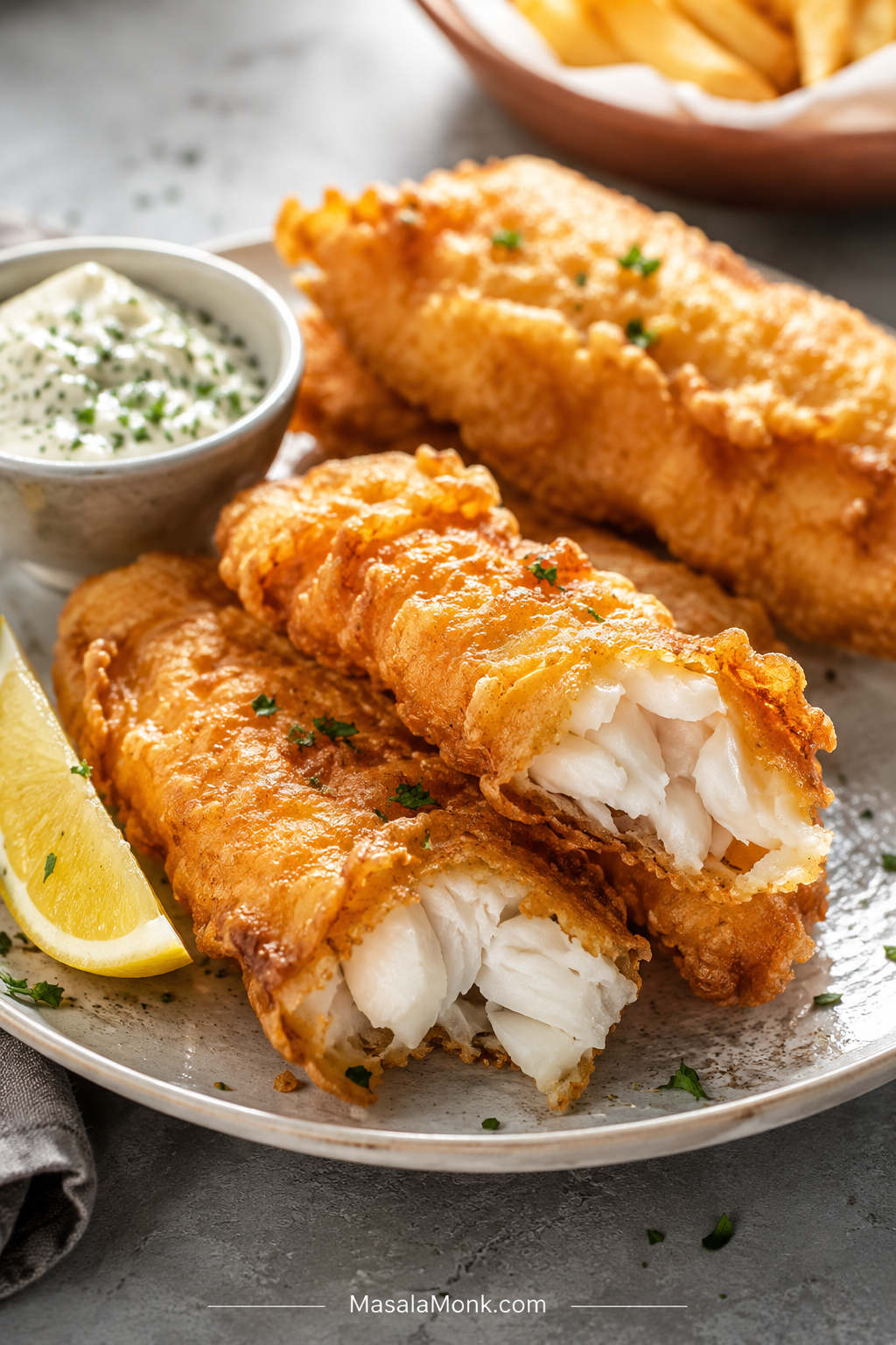 Golden battered fish pieces on a plate with one piece broken open to show flaky white fish, served with lemon and dipping sauce