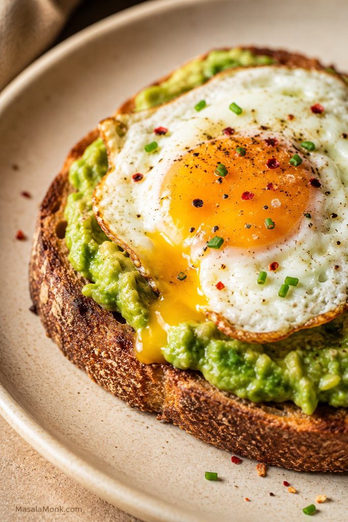 Close-up fried egg avocado toast with a soft yolk, seasoned avocado, chili flakes, and crisp bread crust.
