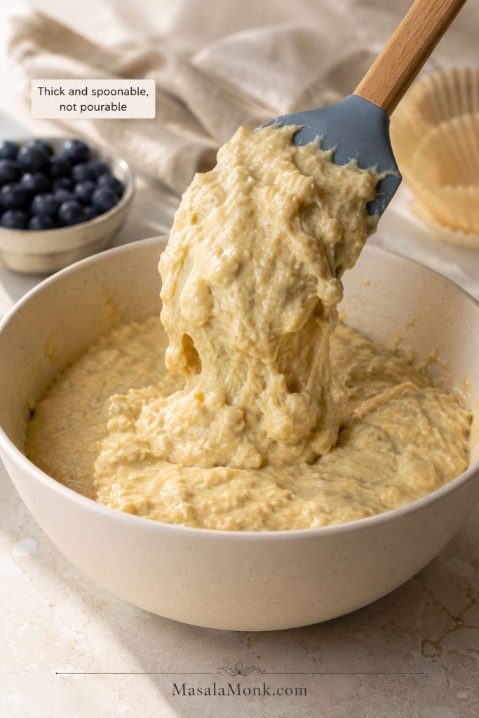 Thick blueberry muffin batter lifted on a spatula with blueberries nearby but not mixed in yet.