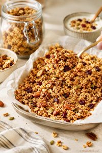 Golden homemade granola with oats, nuts, seeds, and clusters being lifted with a spoon, with a jar and yogurt bowl in the background.