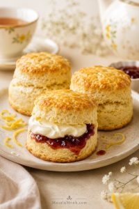 Tall golden lemonade scones on a light tea table, with one scone split open and filled with jam and cream beside a teacup.